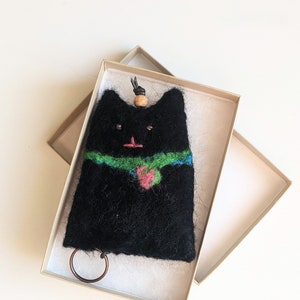 May include: A black felt cat keychain with a green and blue scarf and a pink flower. The keychain is in a small box.
