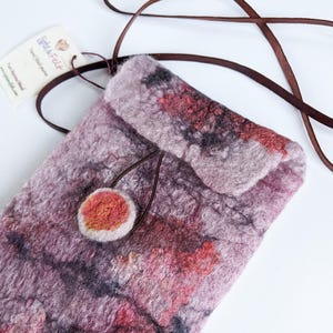 May include: A small, handmade felted bag with a brown leather strap. The bag has a unique, abstract design with shades of pink, red, and gray. The bag has a small, round, felted button.