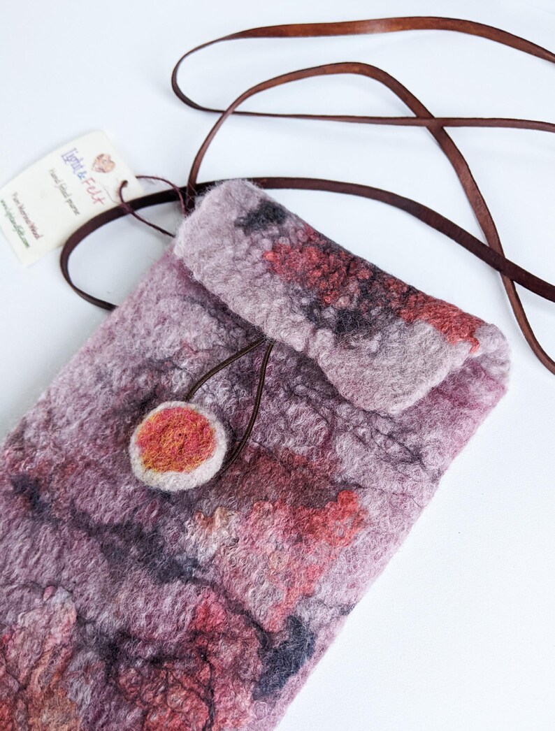 May include: A small, handmade felted bag with a brown leather strap. The bag has a unique, abstract design with shades of pink, red, and gray. The bag has a small, round, felted button.