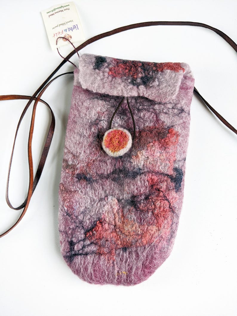 May include: A small, felted bag with a pink, red, and black marbled design. The bag has a brown leather strap and a round, orange button closure. The bag is handmade and has a unique, artistic look.
