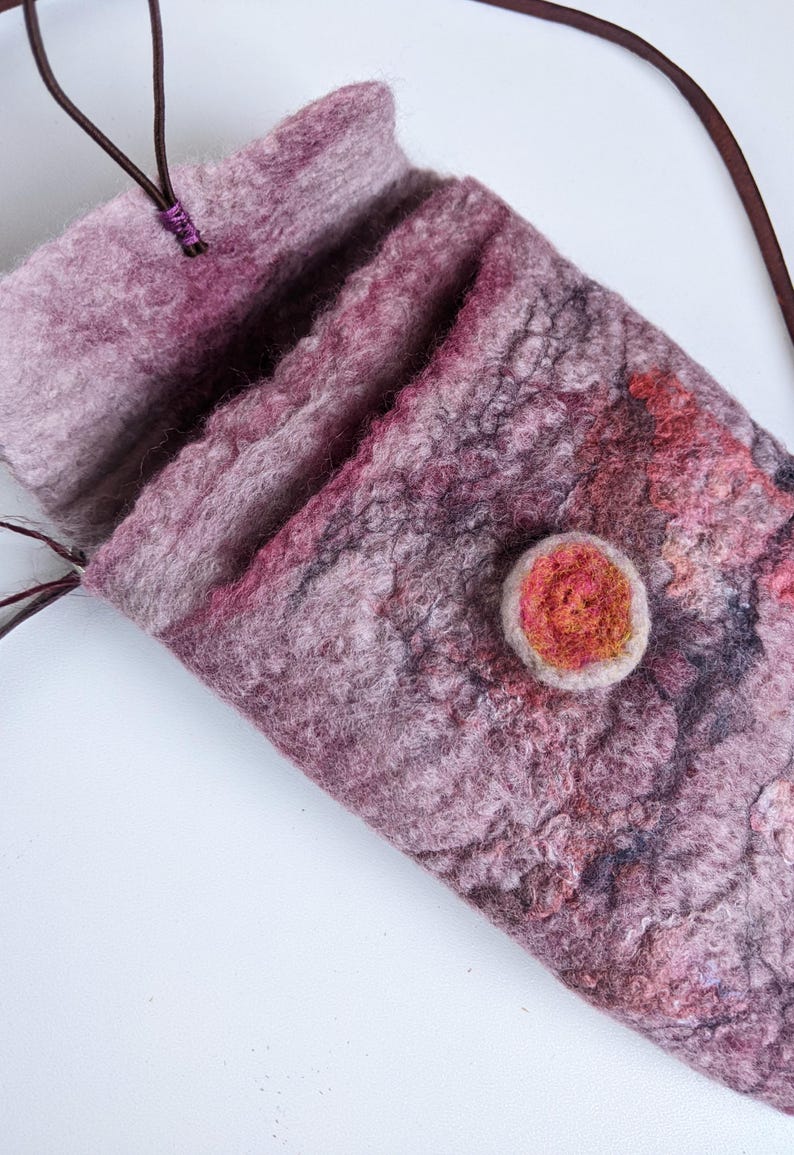 May include: A small, pink and purple felted bag with a brown leather strap and a red felt button.