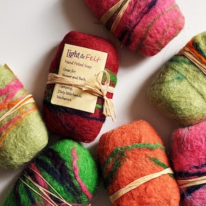 May include: A close-up of several colorful felted soap bars. The soap bars are wrapped in twine and have a variety of colors, including green, red, orange, and pink. The soap bars are labeled "Light & Felt" and are described as "Hand Felted Soap".