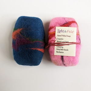May include: Two hand felted soap bars, one blue and one pink, with colorful wool accents. The pink bar has a label that reads "Light & Felt Hand Felted Soap Great for: Shower and Bath Camping, Dirty little hands Mechanics".