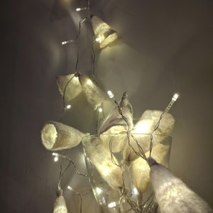 May include: White felt bell-shaped ornaments strung on a strand of fairy lights in a clear glass vase.