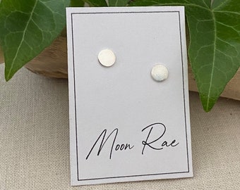 May include: A pair of small, round, silver earrings on a white card with the text "Moon Rae" written in black.