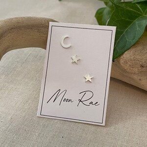 May include: A set of three silver earrings featuring a crescent moon and two stars. The earrings are displayed on a white card with the text "Moon Rae".