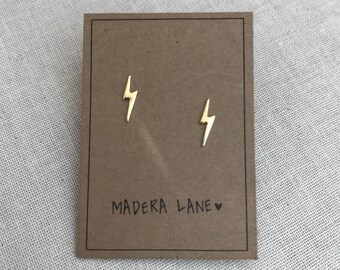 Puede incluir: Un par de pendientes de botón dorados con forma de rayo, presentados en una tarjeta marrón. La tarjeta tiene el texto "MADERA LANE" impreso en negro. Los pendientes son pequeños y minimalistas.