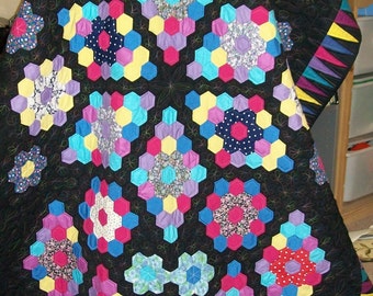 May include: A black quilt with a hexagonal pattern. Each hexagon is made of different fabrics with floral prints in shades of pink, blue, yellow, and purple. The quilt is quilted with a black thread.