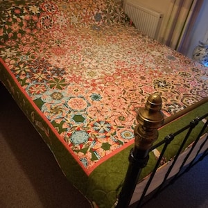 Op de afbeelding: Een kleurrijk patchwork-deken met een groene rand, getoond op een zwart metalen bedframe.