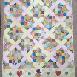 May include: A patchwork quilt with a yellow, pink, and blue color scheme. The quilt is made up of small squares of fabric sewn together in a pattern. The quilt has a border of pink and yellow fabric. The quilt is decorated with a small, embroidered figure of a girl in a pink dress and a blue hat.