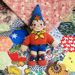 Puede incluir: Un juguete de peluche rojo y azul de un niño que lleva un sombrero azul y una bufanda amarilla con lunares rojos. El juguete está sentado en una colcha de retazos colorida.