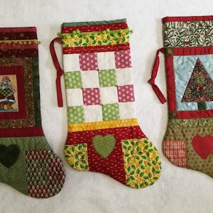 May include: Three handmade Christmas stockings with different designs. The first stocking has a red, green and brown patchwork design with a small image of Santa Claus. The second stocking has a red, green, white and yellow patchwork design with a green heart. The third stocking has a red, green and blue patchwork design with a green Christmas tree.