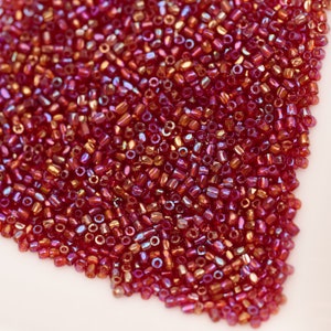 May include: A close-up of a large quantity of small, red iridescent glass beads. The beads are arranged in a loose pile, creating a textured surface.