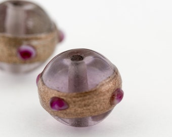 Op de afbeelding: Twee ronde glazen kralen met een gouden band en roze accenten. De kralen zijn doorschijnend en hebben een lichte paarse tint.