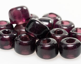 May include: A collection of small, barrel-shaped beads in a deep, translucent purple hue. Each bead features a smooth, glossy finish and a central hole for stringing, with slight variations in shape and color intensity across the group.
