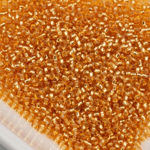 May include: Close-up of a pile of small, golden-colored glass beads. The beads are cylindrical and have a shiny, reflective surface. The beads are densely packed together, creating a textured surface. The beads are likely used for jewelry making or other craft projects.