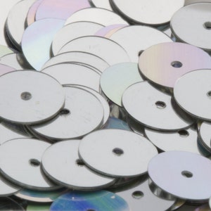 May include: A pile of silver and iridescent round sequins with a hole in the center. The sequins are scattered randomly and some are overlapping.