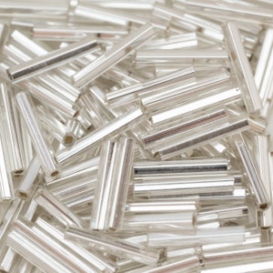 Könnte beinhalten: Nahaufnahme eines Stapels silberner Glasperlen. Die Perlen sind lang und dünn und haben eine reflektierende Oberfläche.