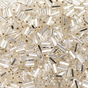 Op de afbeelding: Een close-up van een stapel heldere glazen kralen met een zilveren afwerking. De kralen zijn klein en cilindrisch van vorm.