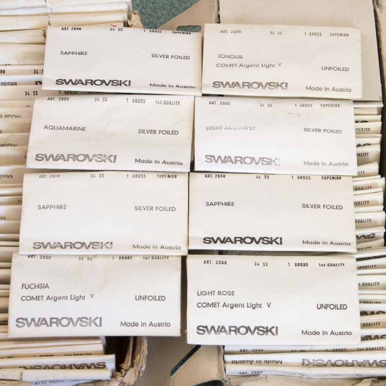 Puede incluir: Primer plano de varias cajas peque&ntilde;as blancas con el texto "SWAROVSKI" impreso en ellas. Cada caja tiene un nombre de color diferente y una descripci&oacute;n del producto, como "SAPPHIRE" o "LIGHT ROSE".