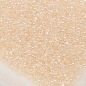 May include: A close-up view of a pile of small, translucent, pale peach-colored seed beads. The beads are round with a small hole in the center, and they are densely packed together. The beads have a slightly iridescent quality.
