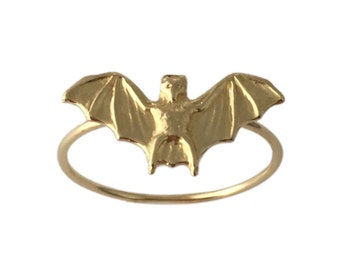 May include: A gold-colored ring with a bat design. The bat is in a flying position with its wings spread out.