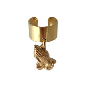 May include: Gold ear cuff with a dangling praying hands charm. The cuff is a simple band with a rounded edge. The charm is detailed and textured.