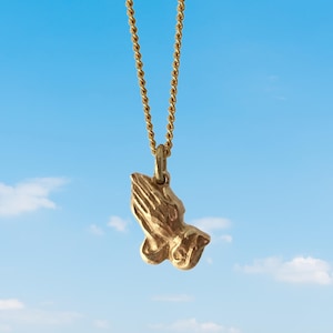 May include: A gold chain necklace with a praying hands pendant. The pendant is a detailed, three-dimensional sculpture of two hands clasped in prayer.