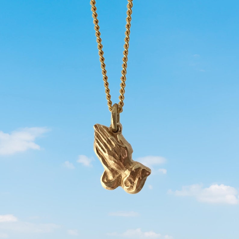 May include: A gold chain necklace with a praying hands pendant. The pendant is a detailed, three-dimensional sculpture of two hands clasped in prayer.