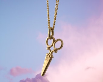 May include: A gold chain necklace with a gold scissor pendant. The pendant is detailed and has a vintage look. The necklace is hanging against a blue and pink cloudy sky.