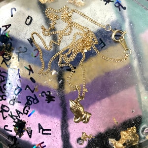 May include: A gold chain necklace with a praying hands pendant. The necklace is on a clear surface with gold flecks.