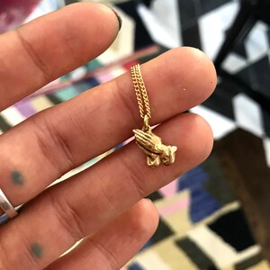 May include: A gold chain necklace with a small gold pendant in the shape of praying hands.