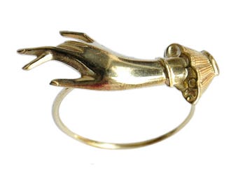 May include: A gold-toned ring with a sculpted hand design. The hand is reaching out with the fingers extended. The ring is made of a thin metal band.