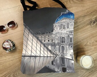 May include: Black tote bag featuring a grayscale painting of the Louvre Museum in Paris, France, with a blue dome. The bag has black straps. Sunglasses and a decorative bracelet are next to the bag.