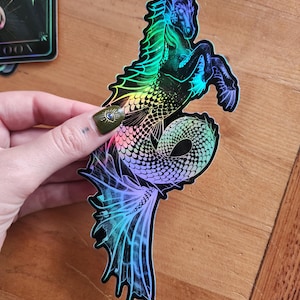 May include: A holographic sticker featuring a mythical sea creature with the head and front legs of a horse and a long, scaled fish tail. The design displays a vibrant spectrum of iridescent colours including blue, green, and purple against a black background.