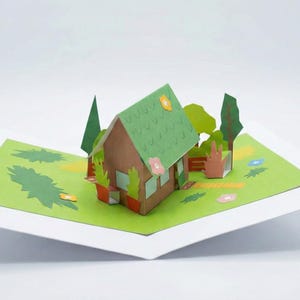Peut inclure: Une carte pop-up représentant une maison marron avec un toit vert et une scène de jardin. La maison a de petites fenêtres et est entourée d'arbres verts et de fleurs sur une pelouse verte. La carte est ouverte, révélant l'art en papier 3D.