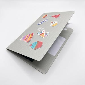 May include: A gray notebook with colorful stickers of cupcakes, glasses, and other designs. The notebook is open, revealing a blank white page. The stickers are in various colors, including red, blue, yellow, and pink.