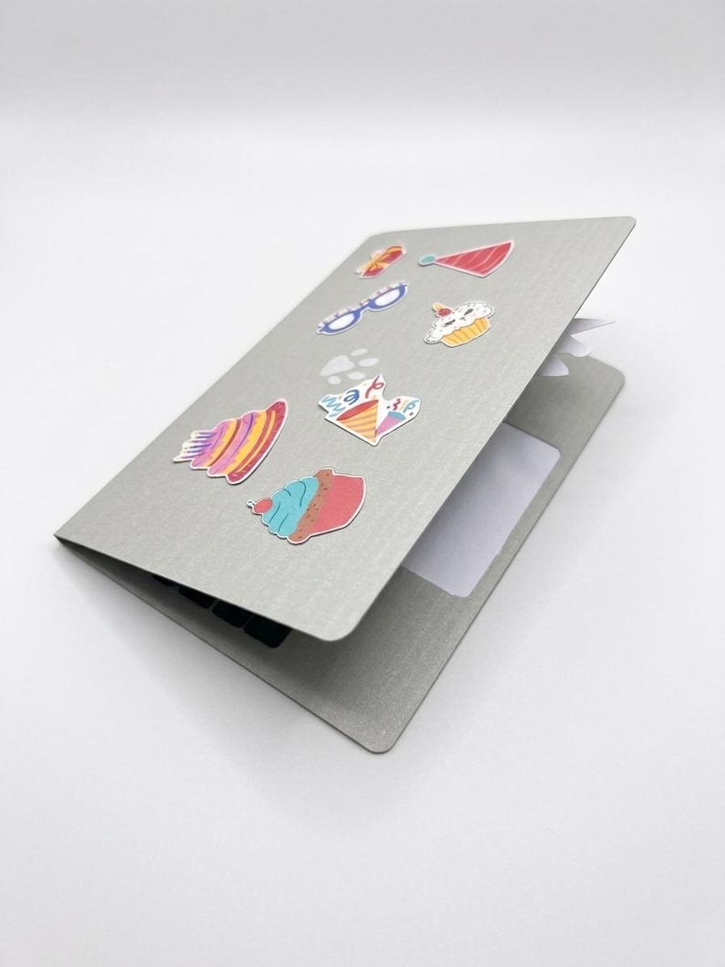 May include: A gray notebook with colorful stickers of cupcakes, glasses, and other designs. The notebook is open, revealing a blank white page. The stickers are in various colors, including red, blue, yellow, and pink.