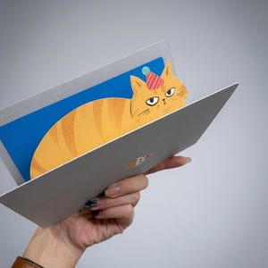 May include: A greeting card featuring a cartoon orange tabby cat wearing a party hat. The cat is inside a laptop-shaped card with a blue screen. The card is held against a gray background.