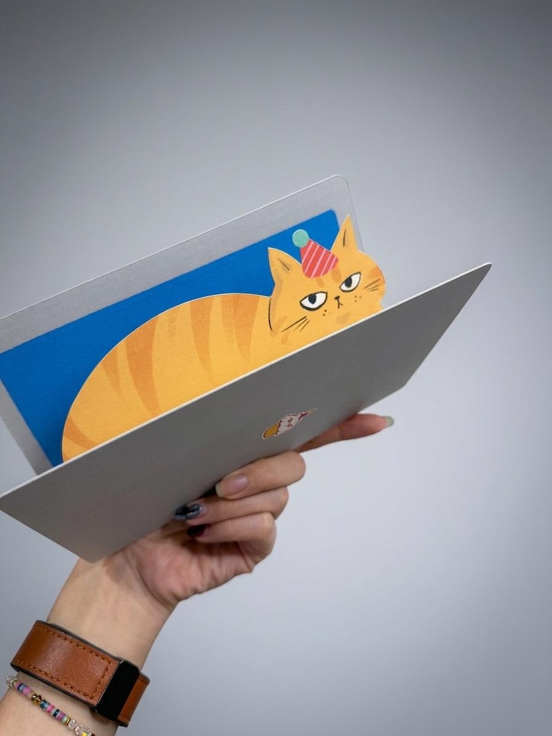 May include: A greeting card featuring a cartoon orange tabby cat wearing a party hat. The cat is inside a laptop-shaped card with a blue screen. The card is held against a gray background.