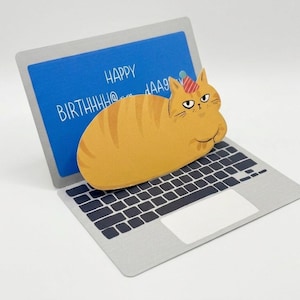 May include: A birthday card featuring a cartoon orange tabby cat wearing a party hat, perched on a laptop. The laptop screen displays the words "HAPPY BIRTHHHH@..." in white against a blue background. The card is on a white surface.