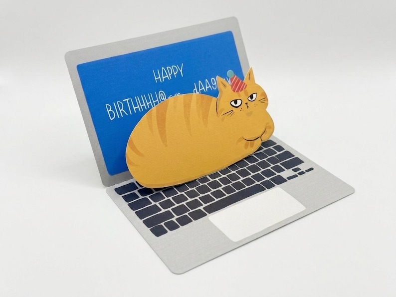 May include: A birthday card featuring a cartoon orange tabby cat wearing a party hat, perched on a laptop. The laptop screen displays the words "HAPPY BIRTHHHH@..." in white against a blue background. The card is on a white surface.