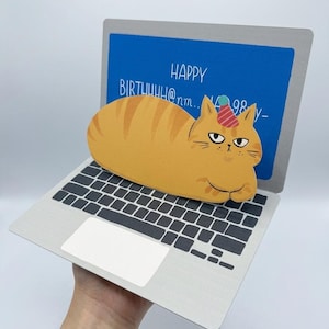 May include: A whimsical birthday card shaped like a laptop, featuring a cartoon orange tabby cat wearing a party hat. The laptop screen displays "HAPPY BIRTHUUH@nm... 98 y-". The keyboard and trackpad are visible.