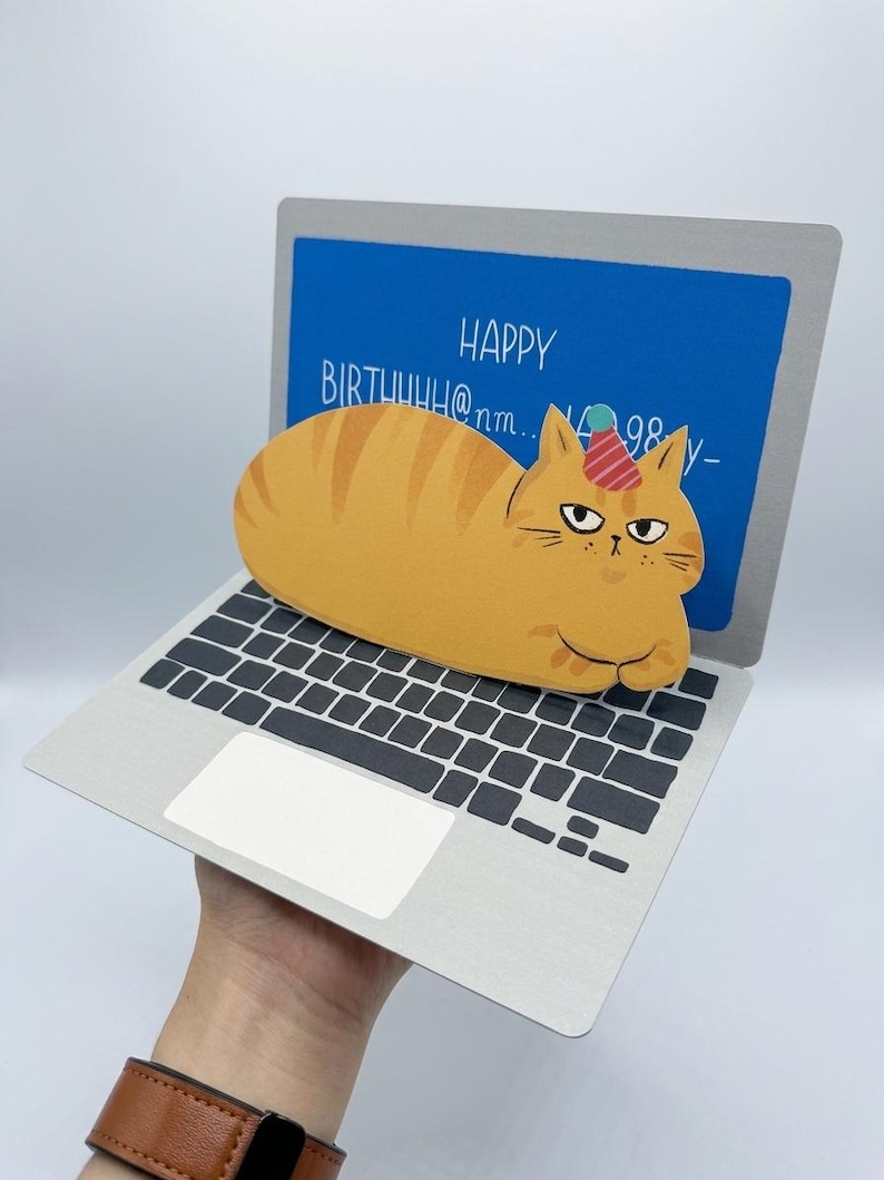 May include: A whimsical birthday card shaped like a laptop, featuring a cartoon orange tabby cat wearing a party hat. The laptop screen displays "HAPPY BIRTHUUH@nm... 98 y-". The keyboard and trackpad are visible.
