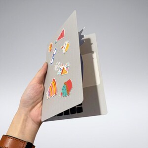 May include: A gray laptop with colorful stickers, held in a hand with a brown leather watch. The stickers include images of a red party hat, glasses, and various food items. The laptop's keyboard is visible.