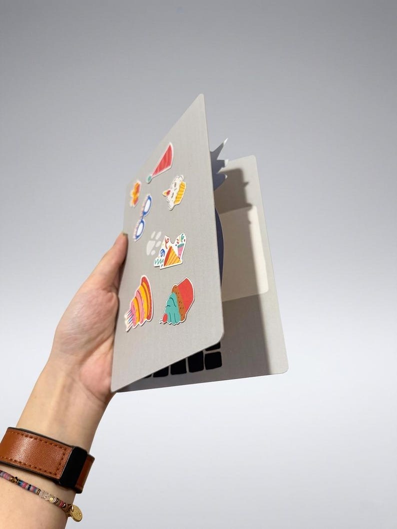 May include: A gray laptop with colorful stickers, held in a hand with a brown leather watch. The stickers include images of a red party hat, glasses, and various food items. The laptop's keyboard is visible.