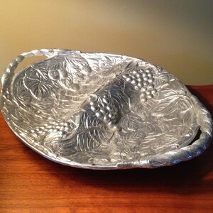May include: Silver-colored, oval serving dish with a grape and leaf design. The dish has two sections and a braided handle on each end. The detailed relief pattern is visible on the interior and exterior of the dish.