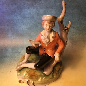 May include: A porcelain figurine depicting a seated man in an orange jacket, black breeches, and a white ruffled shirt, holding a small bird. The figure is posed near a tree trunk, with a floral base. The figurine has a vintage aesthetic.