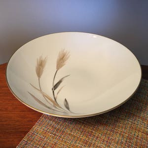 May include: A cream-colored, round serving bowl with a gold rim. The bowl features a wheat stalk design in shades of brown, gray, and black. The bowl is set on a wooden surface.