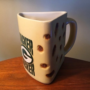 May include: A cream-colored, triangular mug with a handle, featuring the words "PACKER" in green and a green and white logo. The mug has brown, irregular holes, resembling cheese. The mug is on a wooden surface.